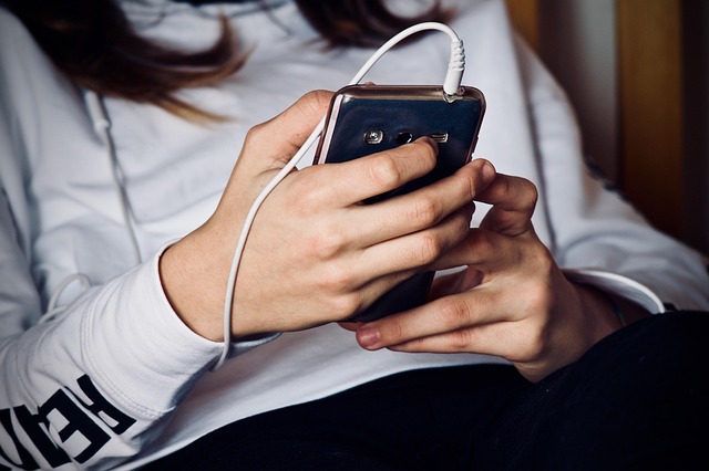 En ung person i grå tröja sitter och scrollar på sin mobil. Huvudet syns inte.