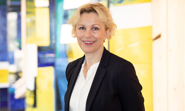 Maria Strandberg i vit blus och mörk kavaj framför en ljus vägg med konstverk i blått och gult.