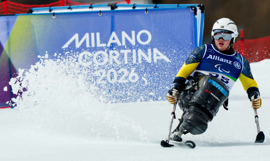 Elijah åker sitski. Snö sprutar från skidan. De tstår Milano Cortina 2026 i bakgrunden. Han ha r vit hjälm och blågul dress.