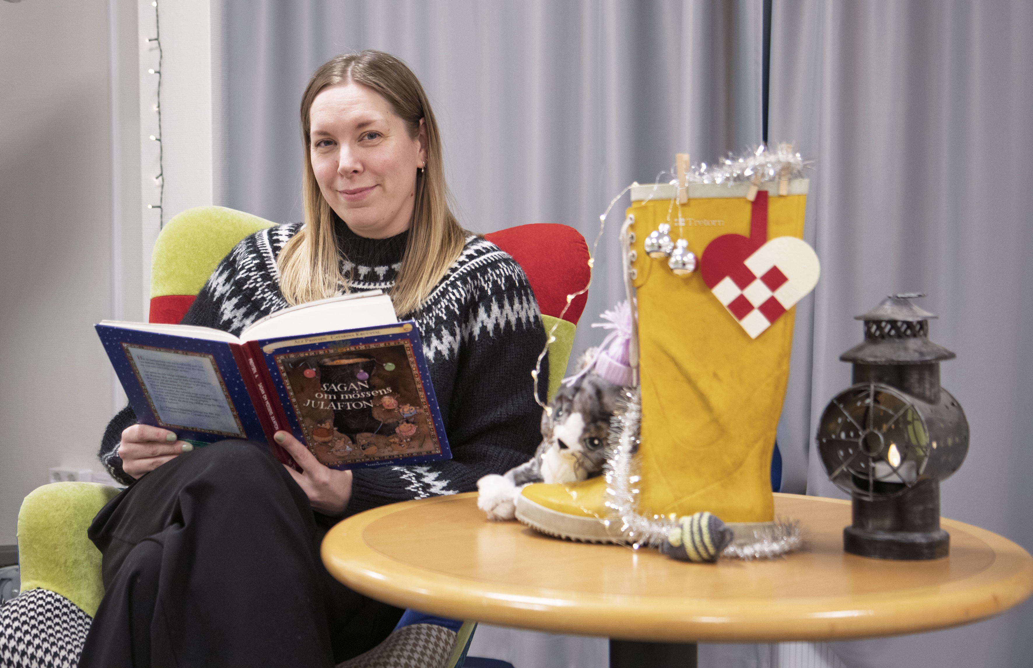 Monica Korp sitter vid ett bord och läser ur en bok. Olika rekvisita på ett bord bredvid.