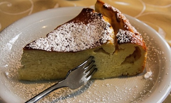 En bit cheesecake på assiett med en gaffel instucken i den. Florsocker pudrat över.