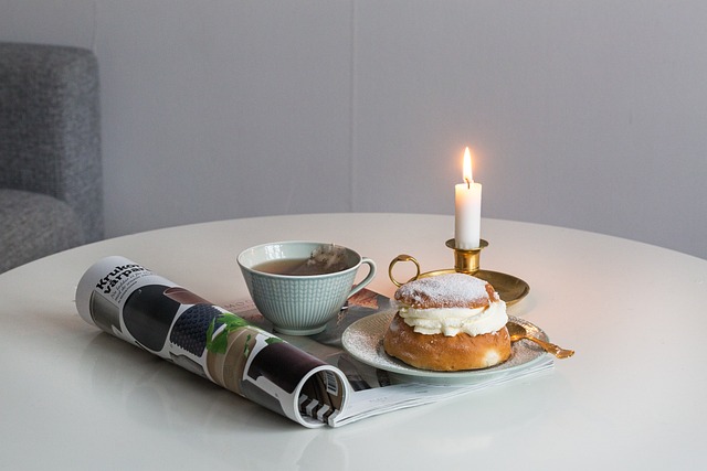 Ett vitt bord med en tidning uppslagen. Det står en semla och en kopp kaffe på bordet. Bredvid är det ett tänt ljus.