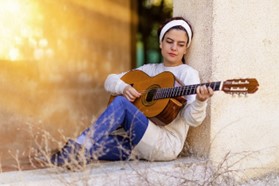 En tjej sitter lutad mot en husvägg och spelar på en gitarr. Hon har vit tunika och vitt hårband. Solen speglar sig i fönstret bredvid.