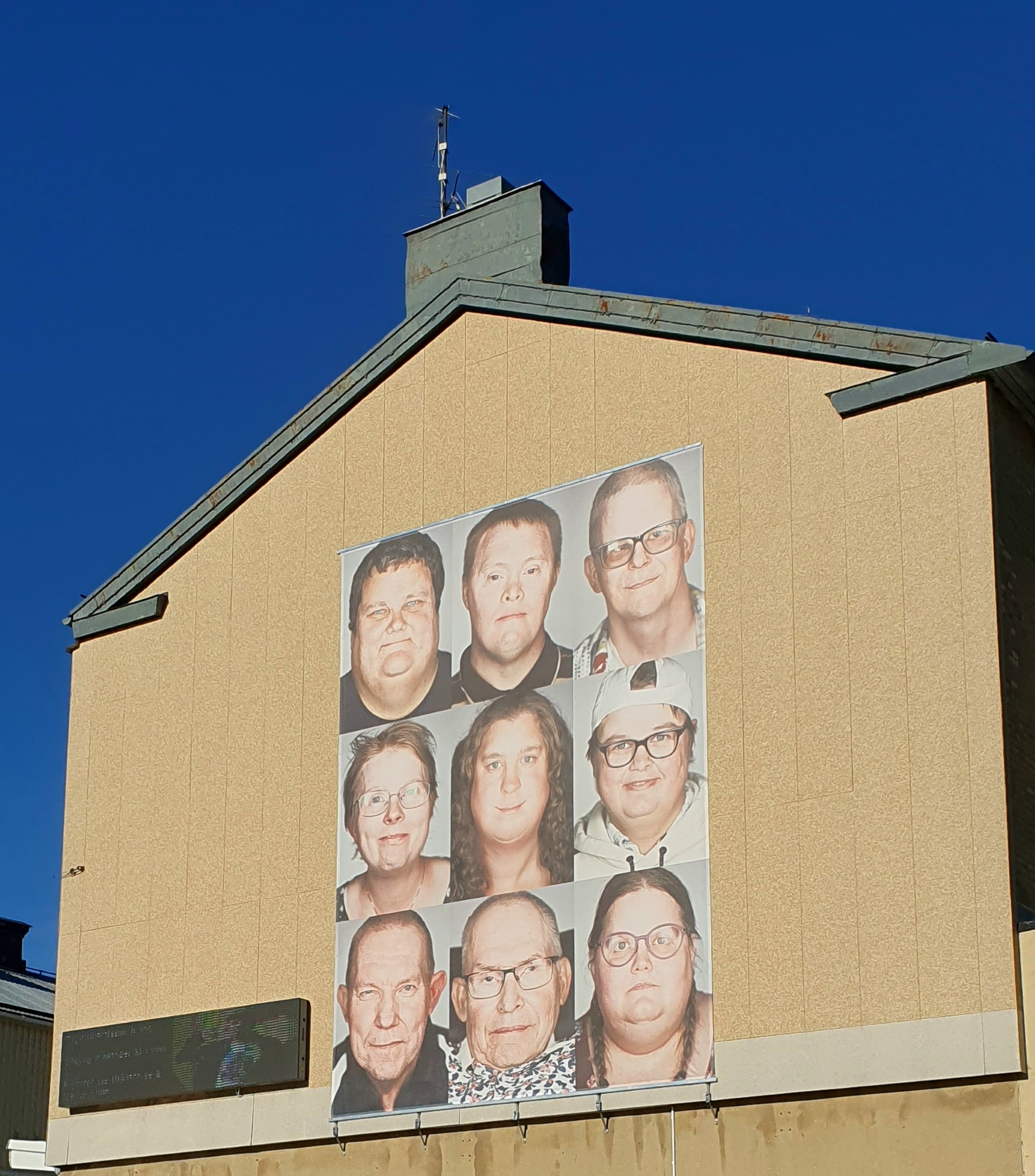 Stor plansch på husvägg. Ansikten på nio personer med intellektuell funktionsnedsättning