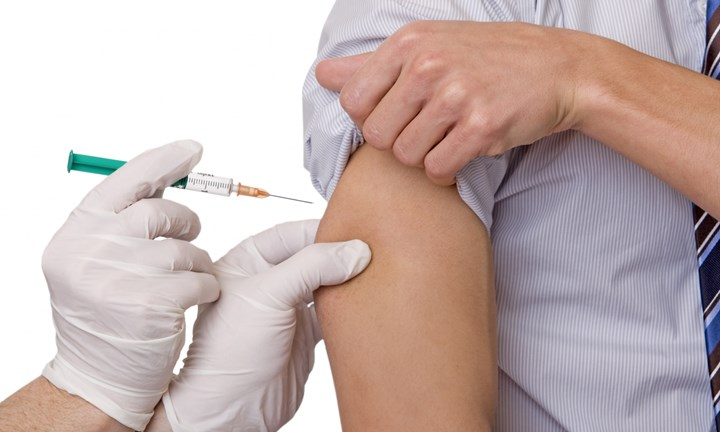 Närbild på överarm. Personen håller i den upprullade skjortärmen. Personen får en  vaccination i överarmen. Handskklädda händer håller i sprutan.