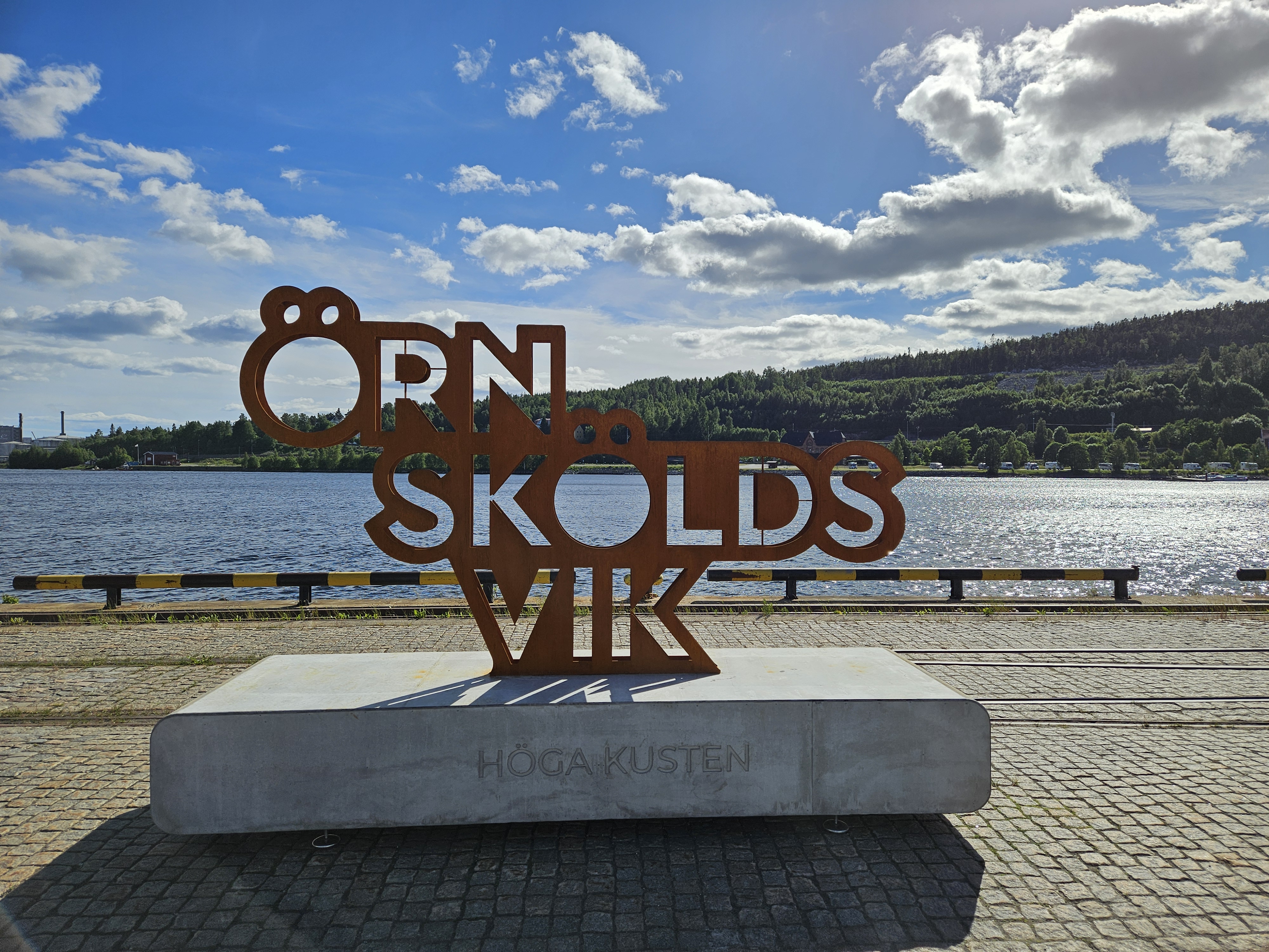 Nya landmärket vid kajen, med blå himmel och glitter i vattnet. Örnsköldsvik i stora rostbruna bokstäver på en stor grå cementunderdel. Den går att stå och sitta på.