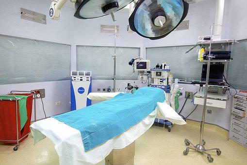 En operationsal som står redo att ta emot patient. Operationssäng i mitten med dropställning bredvid. Väggarna är ljusgrå och gröna. Medicinsk apparatur på bänk och en rullvagn. Stora operationslampor i taket.