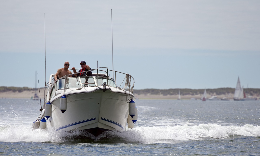 motorbåt sedd framifrån. Det skummar och är lite vågor runtom. segelbåtar och en strand i bakgrunden. Blå himmel.