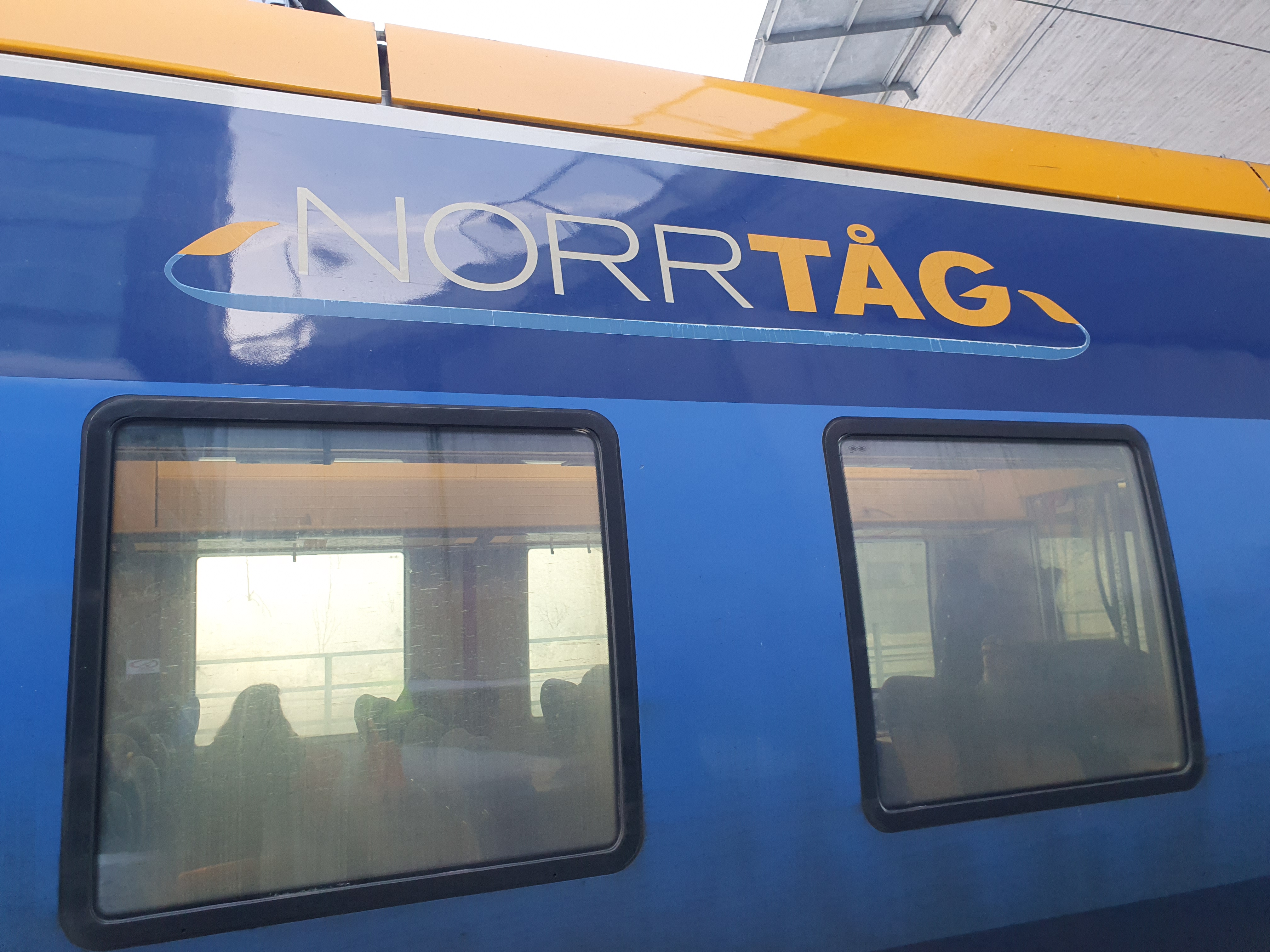 Närbild på Norrtågs vagn i två nyanser av blått. Det står Norrtåg i brandgult och vitt ovanför fönstren. Det går att skymta passagerare genom fönstren.