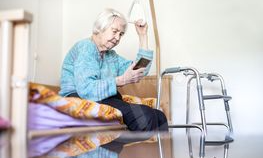 Äldre kvinna sitter på sjukhussäng och håller i sig i däverten. Hon har mobiltelefon i andra handen. Rullatorn stå framför henne.