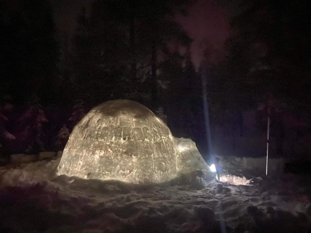 En igloo med en lampa som lyser in genom ingången . Det ser ut som en stor snölykta i kvällsmörkret.