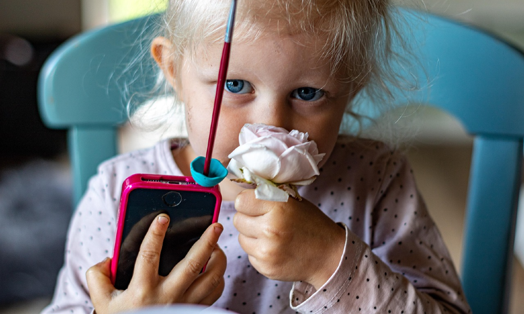 Yngre barn med mobiltelefon i ena handen. Luktar på en ros som barnet håller i den andra handen.
