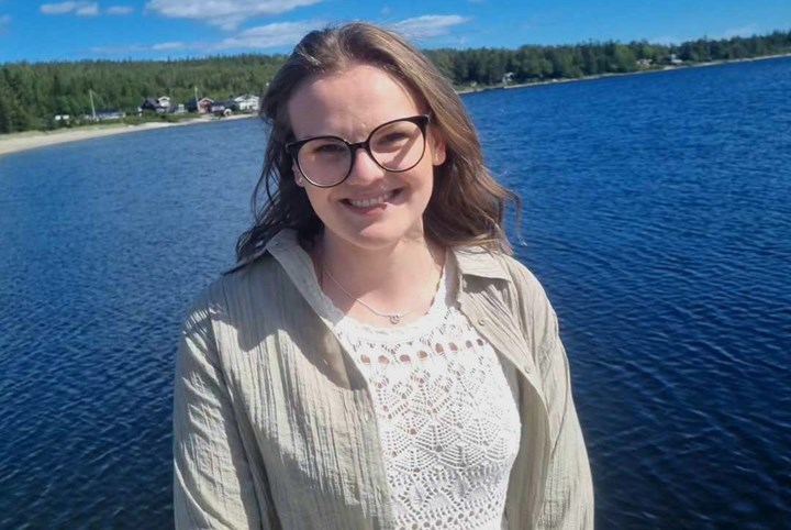 Anna L står utomhus med havet bakom sig. Och det är blå himmel. Hon är glad och håret är lite vindrufsigt. Anna har en ljus tröja med spets. Och en ljus skjorta. Hon har glasögon.