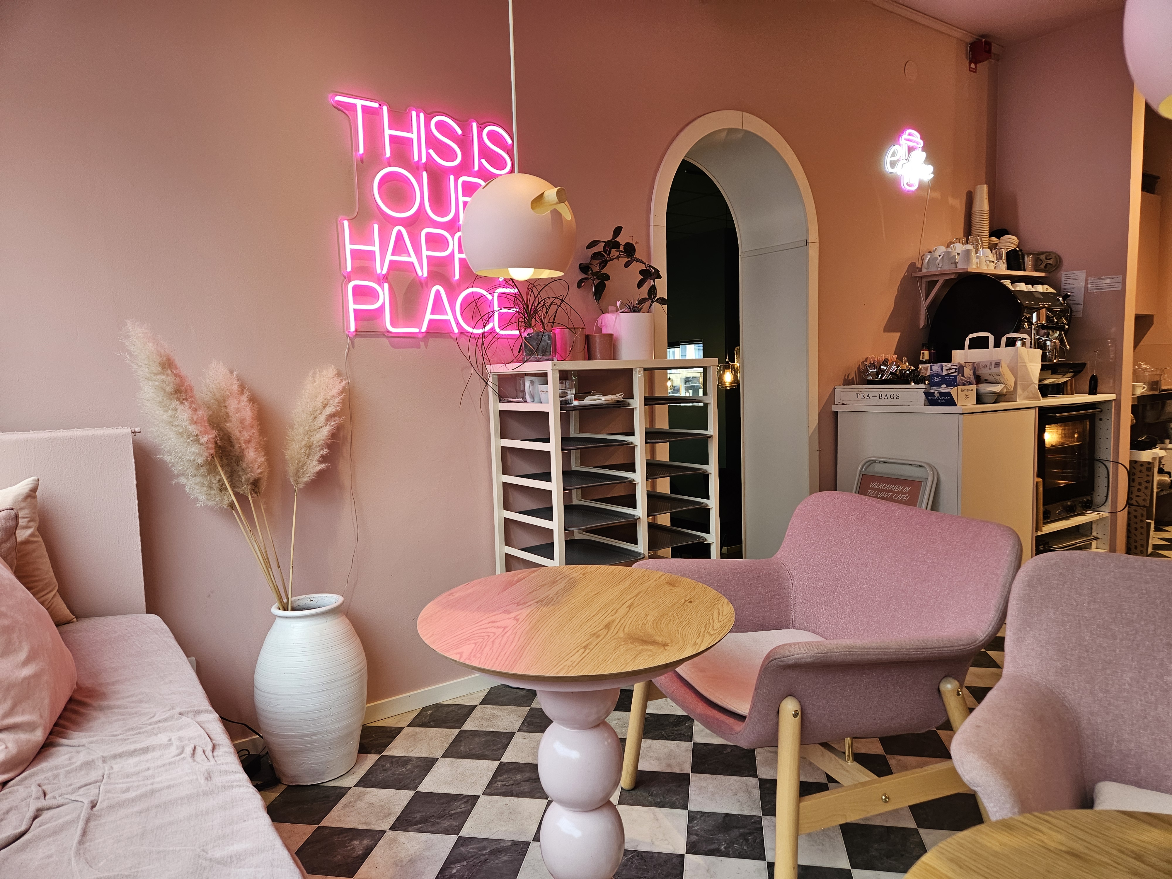Fåtöljer och väggar i rosa. På väggen en upplyst skylt, där det står Our happy place. Shackrutigt golv. Ety ställ för brickor står mot väggen. Bredvid en dörröppning till Hälsobaren.